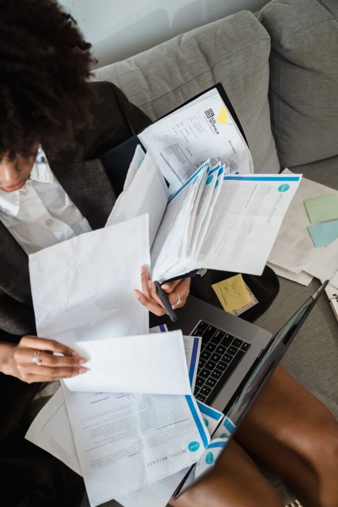 A woman sitting on a couch organizing paperwork with a laptop in front of her.
