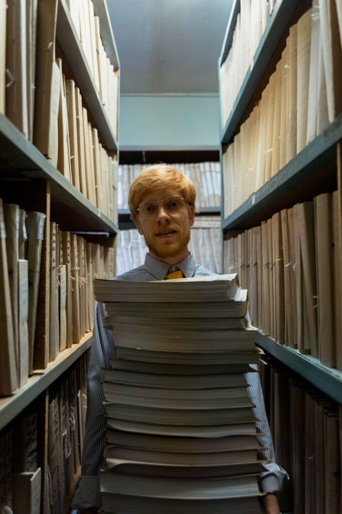Redhead male standing in library aisle, carrying a large stack of documents.